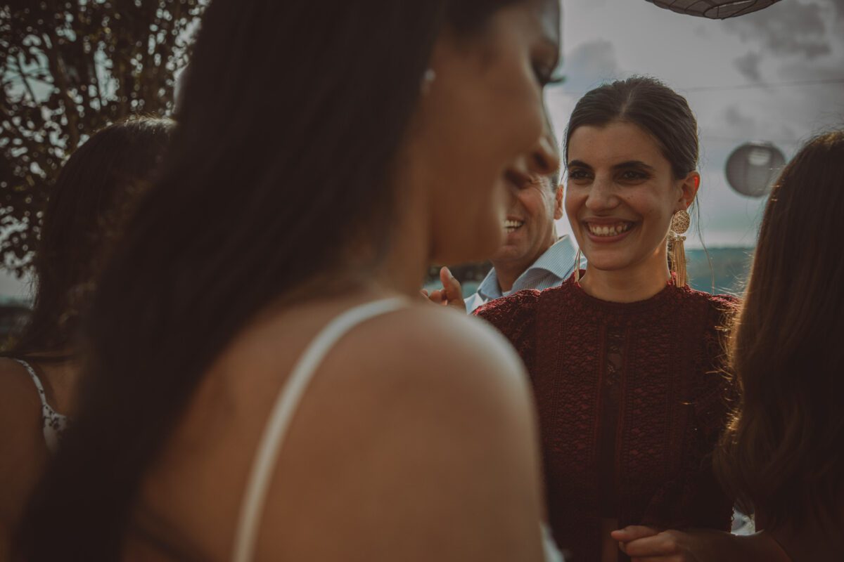 Eine Frau in einem dunkelroten Kleid lächelt strahlend bei einer Versammlung im Freien, möglicherweise einer standesamtlichen Trauung. Sie hat dunkles Haar zurückgekämmt und trägt große, baumelnde Ohrringe. Der Fokus liegt auf ihr, während sie den Moment genießt, während eine andere Frau im Vordergrund vor einem verschwommenen Hintergrund aus Bäumen und Himmel unscharf ist. - Elis Wedding - Hochzeitsfotograf Frankfurt