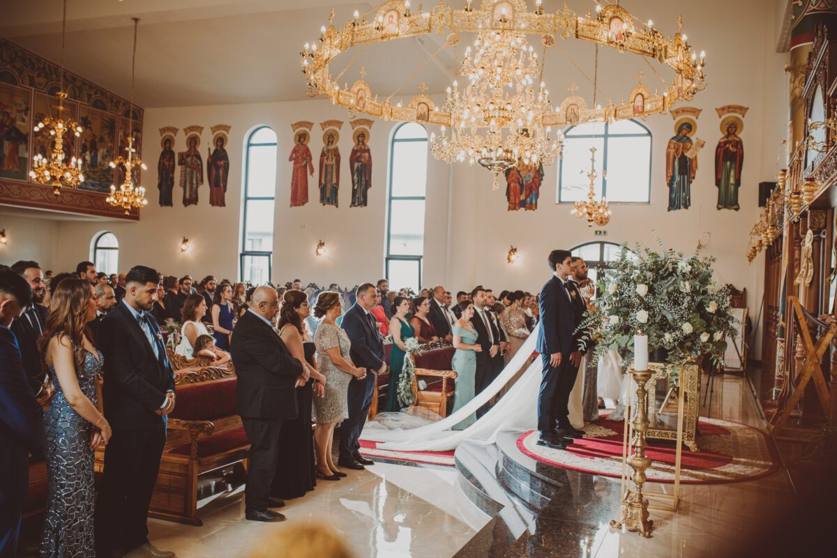 Eine Hochzeitszeremonie findet in einer großen, reich verzierten Kirche in Frankfurt mit hohen Decken und aufwändigen Kronleuchtern statt. Braut und Bräutigam stehen am Altar, ihre lange Schleppe weht hinter ihnen her. Gäste in formeller Kleidung sitzen auf Holzbänken, während ein Hochzeitsfotograf jeden kostbaren Moment inmitten der religiösen Ikonen festhält. - Elis Wedding - Hochzeitsfotograf Frankfurt