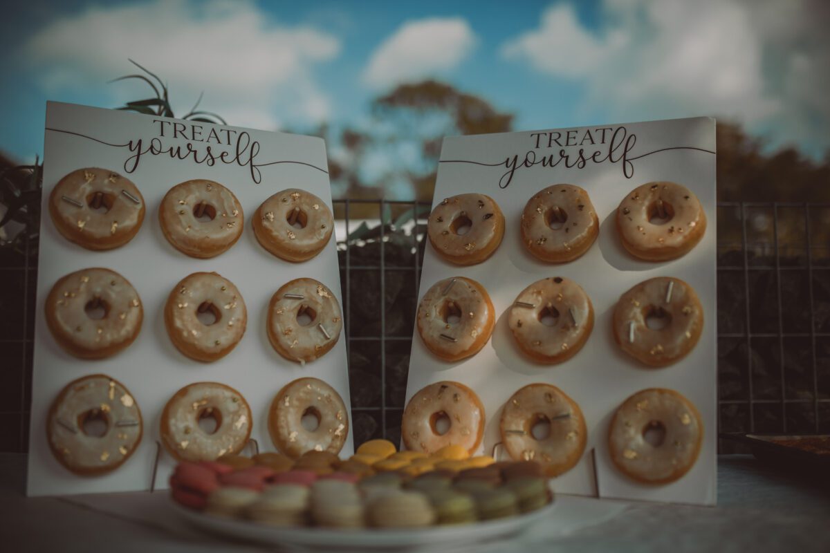 Eine Präsentation der Leckereien umfasst zwei „Treat Yourself“-Bretter, auf denen jeweils neun glasierte Donuts in einem 3x3-Raster angeordnet sind. Vor den Brettern auf einem Tisch steht ein weißer Teller mit bunten Macarons. Der Hintergrund zeigt eine verschwommene Ansicht von Bäumen und einem teilweise bewölkten Himmel, perfekt für ein Fotoshooting durch jeden Hochzeitsfotografen. - Elis Wedding - Hochzeitsfotograf Frankfurt