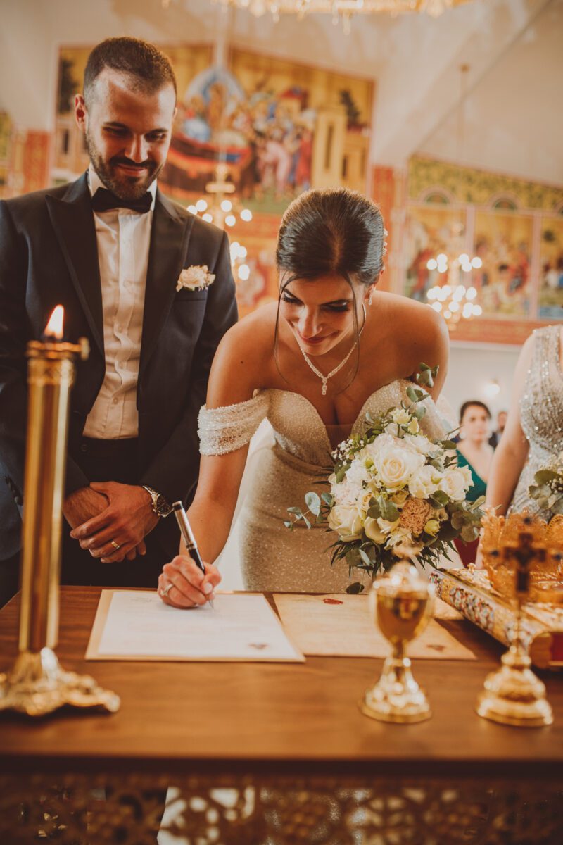 Eine Braut in einem schulterfreien Brautkleid unterschreibt ein Dokument auf einem Holztisch, der mit goldenen Kerzenleuchtern und einem Blumenarrangement geschmückt ist. Der Bräutigam, der einen schwarzen Smoking trägt, steht lächelnd neben ihr. Der Hintergrund zeigt ein lebendiges Dekor mit Kronleuchtern und Gemälden, perfekt für jeden Hochzeitsfotografen, der diesen Moment festhalten möchte. - Elis Wedding - Hochzeitsfotograf Frankfurt