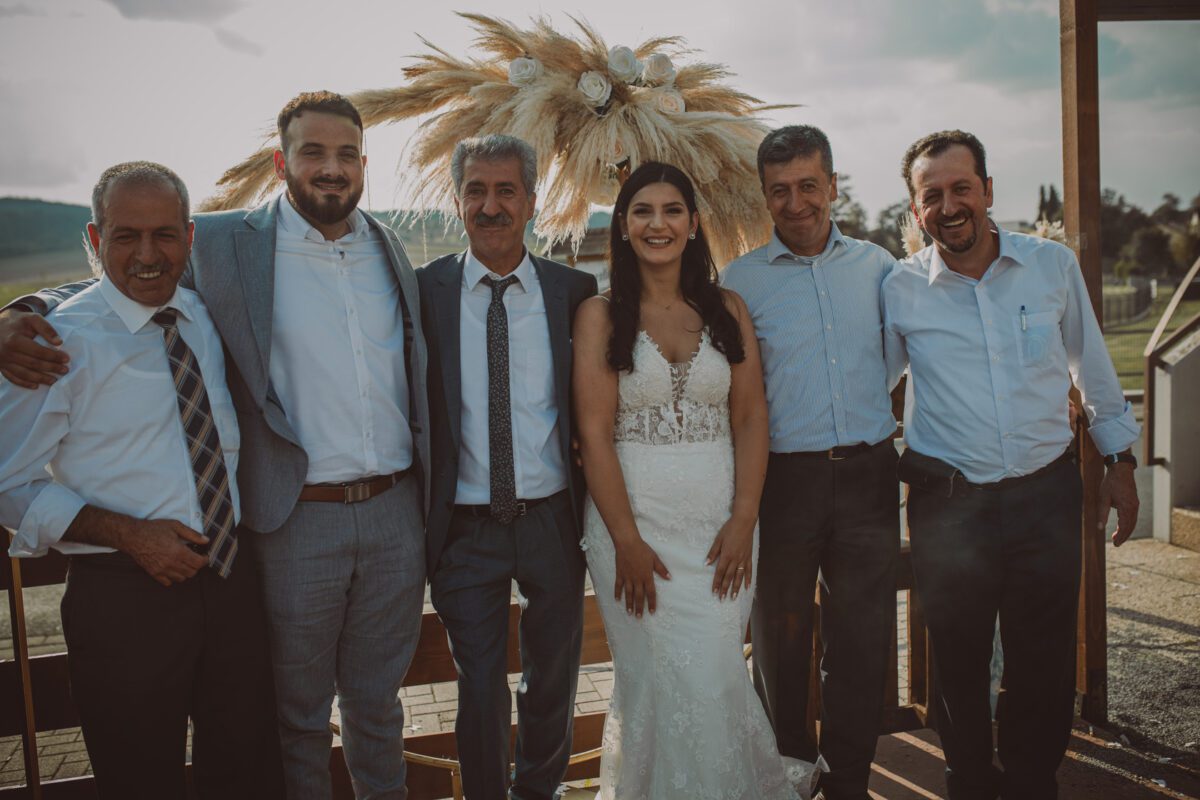 Eine Braut in einem weißen Brautkleid steht lächelnd neben fünf Männern während eines Fotoshootings in Frankfurt. Sie stehen alle im Freien vor einer dekorativen Kulisse aus Pampasgras und Blumen. Die Männer tragen formelle Kleidung, darunter Anzug und Krawatte, und die Umgebung wirkt sonnig und hell. - Elis Wedding - Hochzeitsfotograf Frankfurt