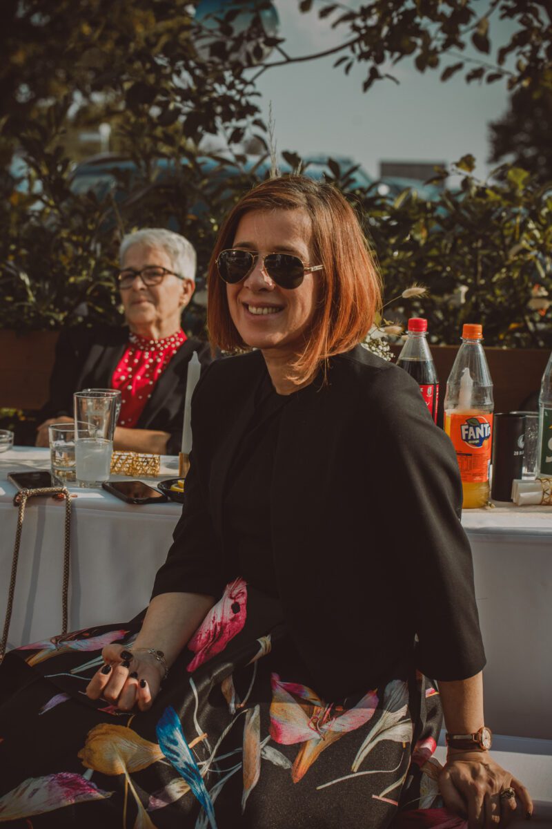 Eine lächelnde Frau mit kurzen, rotbraunen Haaren, einer Sonnenbrille und einem schwarzen Blazer sitzt während eines Fotoshootings in Frankfurt an einem Tisch im Freien mit Getränken und Essen. Hinter ihr sitzt eine ältere Frau mit kurzen, weißen Haaren, einer Brille und einem roten gepunkteten Schal. Im Hintergrund sind Bäume und ein blauer Himmel zu sehen. - Elis Wedding - Hochzeitsfotograf Frankfurt