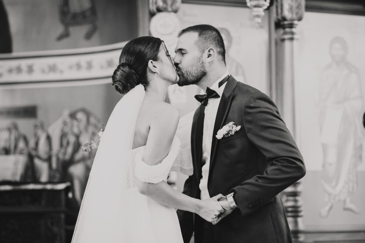 Ein Schwarzweißfoto zeigt Braut und Bräutigam, die sich während ihrer standesamtlichen Trauung küssen. Die Braut in einem trägerlosen Kleid und Schleier und der Bräutigam in einem Anzug mit Fliege und Ansteckblume halten Händchen. Im Hintergrund sind religiöse Symbole und kunstvolle Dekorationen zu sehen – ein Beweis für das Können ihres Hochzeitsfotografen. - Elis Wedding - Hochzeitsfotograf Frankfurt