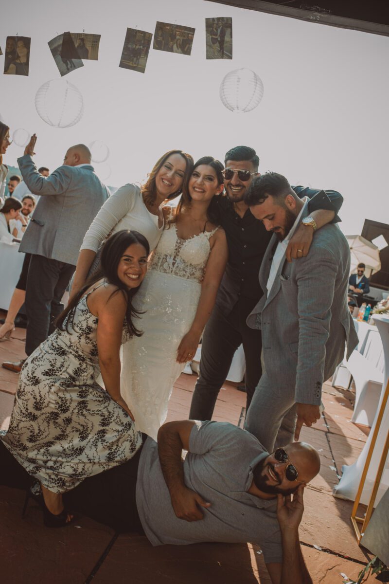 Eine fröhliche Hochzeitsszene mit einer Gruppe von sechs Personen, die zusammen posieren. Die Braut, die ein weißes Kleid trägt, steht in der Mitte, umgeben von lächelnden Freunden in formeller Kleidung. Eine Person liegt verspielt auf dem Boden und trägt zur lebhaften Atmosphäre bei. Der in Frankfurt ansässige Hochzeitsfotograf fängt hängende Fotos und weiße Laternen ein. - Elis Wedding - Hochzeitsfotograf Frankfurt