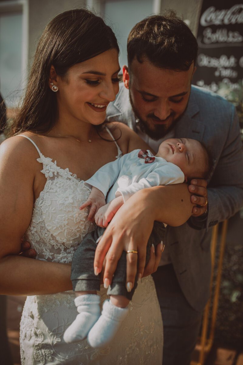 Eine Frau in einem weißen Spitzenkleid lächelt und hält ein schlafendes Baby in den Armen. Ein Mann in einem grauen Jackett steht dicht neben ihr und beugt sich vor, um das Baby mit freudigem Gesichtsausdruck anzusehen. Sie scheinen sich im Freien zu befinden, möglicherweise bei einer Standesamtlichen Trauung in Frankfurt, mit einem Kreidetafel-Schild im Hintergrund. - Elis Wedding - Hochzeitsfotograf Frankfurt