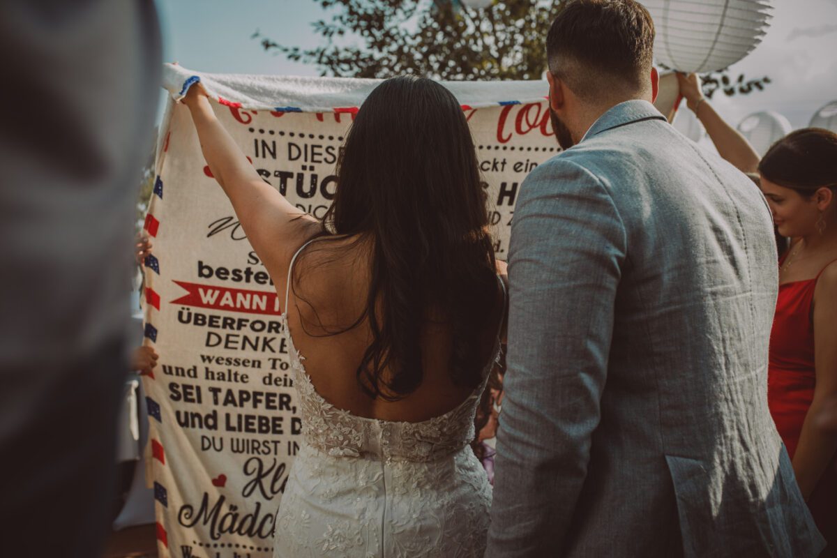 Eine Braut in einem weißen Spitzenkleid mit offenem Rücken und ein Bräutigam in einem hellgrauen Anzug halten bei einer Standesamtlichen Trauung im Freien gemeinsam ein Banner. Die Braut hat langes, dunkles Haar. Das Banner ist mit Text verziert und von Menschen umgeben, von denen einige weiße Laternen halten. Die Kulisse des Fotoshootings wirkt festlich und feierlich. - Elis Wedding - Hochzeitsfotograf Frankfurt