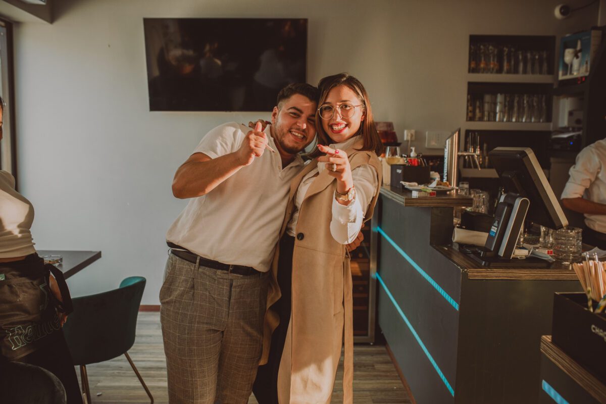 Ein Mann und eine Frau lächeln und zeigen in einer warm beleuchteten Bar in die Kamera, um den Moment ihrer standesamtlichen Trauung festzuhalten. Der Mann trägt ein weißes Hemd und karierte Hosen, während die Frau eine Brille und einen beigen Trenchcoat trägt. Hinter ihnen stehen Flaschen auf der Bartheke unter einem Flachbildfernseher. - Elis Wedding - Hochzeitsfotograf Frankfurt