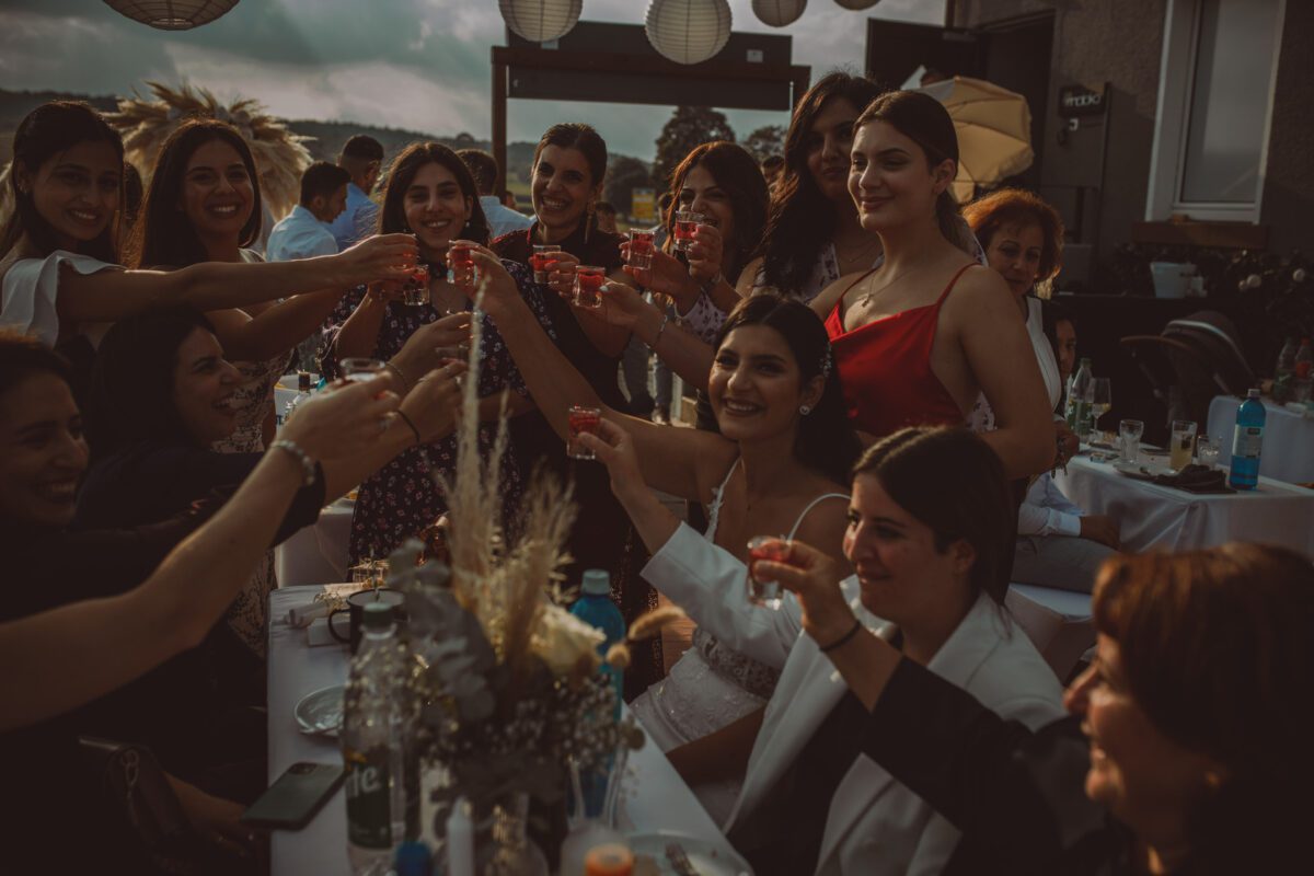 Eine Gruppe von Menschen, hauptsächlich Frauen, sitzt um einen Tisch im Freien in Frankfurt versammelt und stößt zu einem festlichen Toast an. Die Stimmung ist festlich, es wird gelächelt und gelacht. In der Mitte steht eine Frau in einem weißen Kleid, möglicherweise die Braut. Dekorative Laternen hängen darüber und die Umgebung ist warm beleuchtet wie bei einem professionellen Fotoshooting. - Elis Wedding - Hochzeitsfotograf Frankfurt