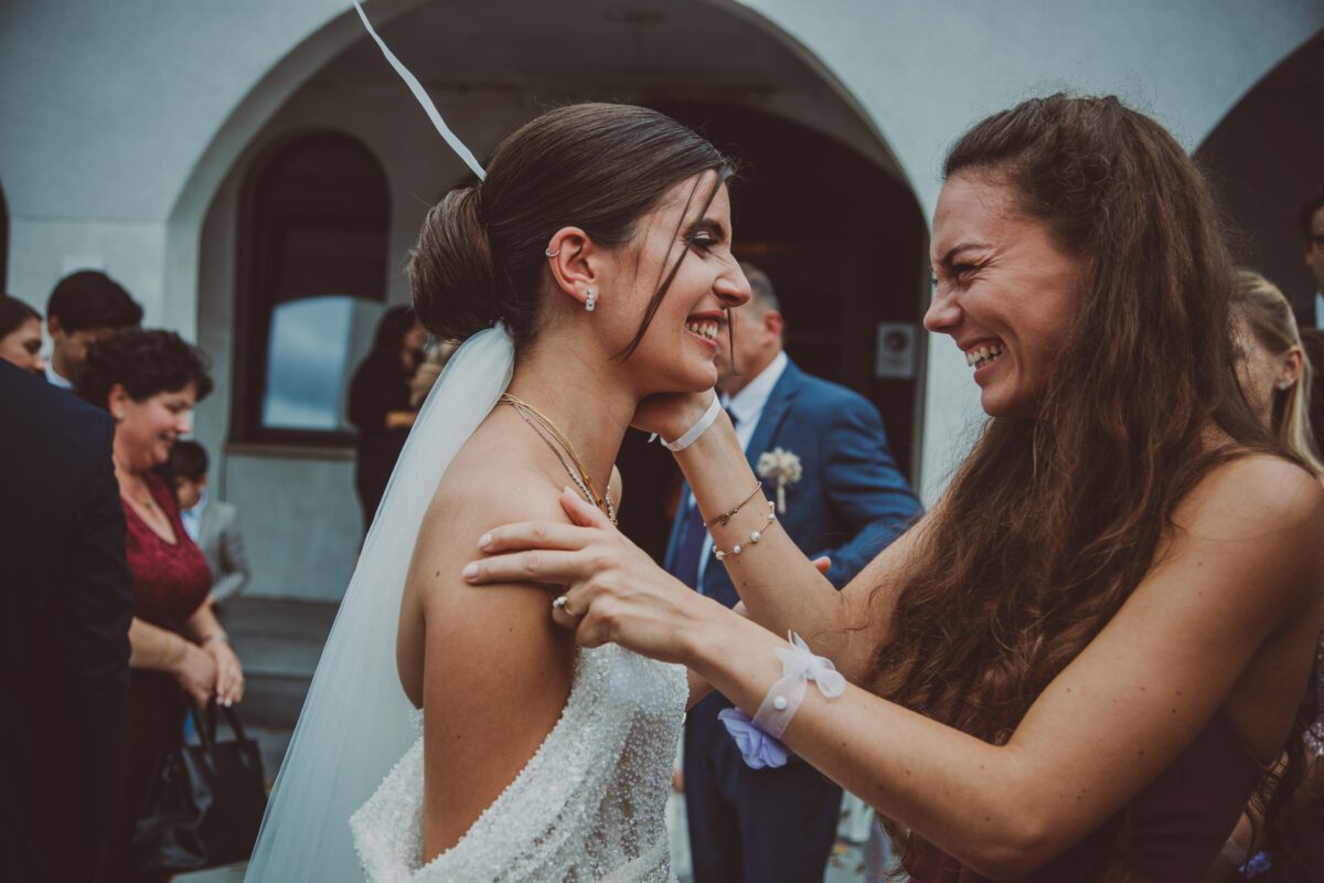 Eine Braut in einem weißen Kleid und Schleier umarmt freudig eine Frau mit langem, offenem Haar während einer Hochzeitszeremonie im Freien. Beide Frauen lachen, und der Hochzeitsfotograf hält jeden kostbaren Moment fest. Sie sind von Zuschauern in formeller Kleidung umgeben und im Hintergrund ist ein gewölbtes Gebäude zu sehen. - Elis Wedding - Hochzeitsfotograf Frankfurt