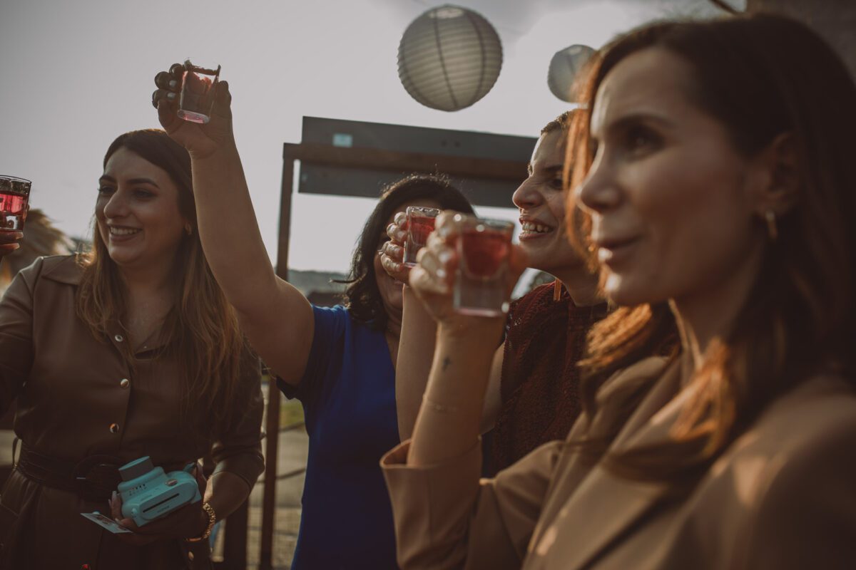 Eine Gruppe von vier Frauen steht dicht beieinander und stößt mit kleinen Gläsern roter Getränke an. Sie stehen vor einem modernen Gebäude mit Papierlaternen und genießen die festliche Atmosphäre einer Standesamtlichen Trauung in Frankfurt. Die Frauen lächeln und scheinen ihre gemeinsame Zeit zu genießen. Sie sind lässig und elegant gekleidet. - Elis Wedding - Hochzeitsfotograf Frankfurt