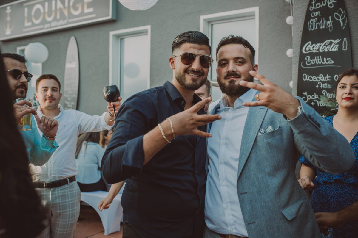 Zwei Männer posieren für ein Foto bei einem Treffen im Freien in Frankfurt. Der Mann links trägt ein dunkelblaues Hemd und eine Sonnenbrille, während der Mann rechts einen hellgrauen Anzug trägt, der vielleicht gerade frisch von einer standesamtlichen Trauung kommt. Beide gestikulieren mit den Händen vor einer grauen Wand mit verschiedenen Schildern. Im Hintergrund unterhalten sich Leute und halten Getränke in der Hand. - Elis Wedding - Hochzeitsfotograf Frankfurt