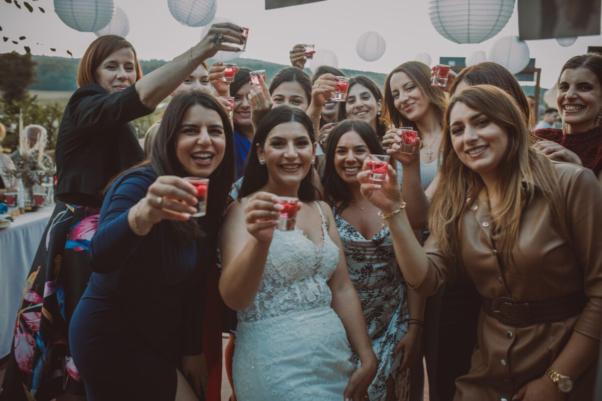 Eine Gruppe von Frauen versammelt sich bei einer Veranstaltung im Freien, lächelt und stößt mit roten Schnapsgläsern an. Die Braut, in ein weißes Kleid gekleidet, steht in der Mitte. Im Hintergrund hängen Papierlaternen und dahinter ist ein Waldstück zu sehen – perfekt, um diesen freudigen Moment während des Fotoshootings festzuhalten. - Elis Wedding - Hochzeitsfotograf Frankfurt