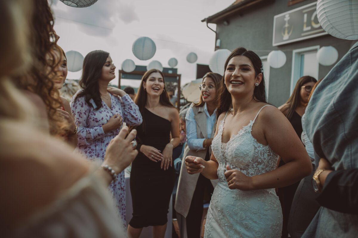 Eine fröhliche Braut in einem weißen Spitzenkleid tanzt während ihrer standesamtlichen Trauung inmitten einer Gruppe von Frauen. Sie befinden sich im Freien in Frankfurt, umgeben von weißen Hängelaternen. Die Frauen lächeln und tragen verschiedene Outfits, darunter Kleider und Jacken. Im Hintergrund ist ein Gebäude mit einem Schild und einem bewölkten Himmel zu sehen, wunderschön eingefangen in Fotografie. - Elis Wedding - Hochzeitsfotograf Frankfurt