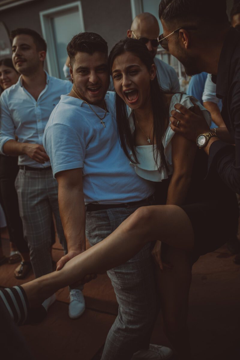 Eine lebhafte Gruppe von Menschen hat Spaß bei einer gesellschaftlichen Veranstaltung. Ein Mann links, der ein weißes Poloshirt trägt, lächelt breit, während er spielerisch das Bein einer Frau hochhebt. Die Frau, in einem weißen Oberteil und einem schwarzen Rock, lacht. Diesen freudigen Moment hält ein leidenschaftlicher Hochzeitsfotograf inmitten der fröhlichen Menge fest. - Elis Wedding - Hochzeitsfotograf Frankfurt