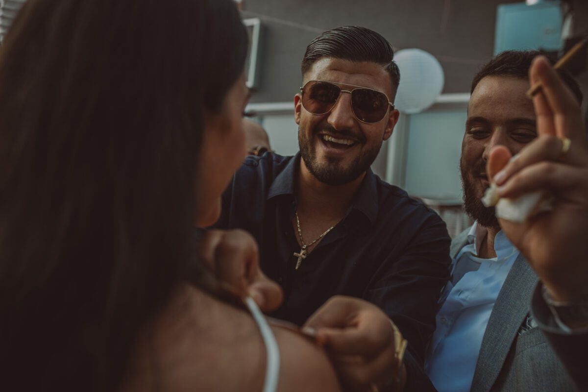 Ein Mann mit Sonnenbrille lächelt, während er einer Frau hilft, eine Nadel an einem Kleiderträger zu befestigen. Ein anderer Mann, der teilweise zu sehen ist, hält eine Zigarre und lächelt ebenfalls. Die Szene scheint ein Fotoshooting im Freien zu sein, mit einem verschwommenen Hintergrund, der Dekorationen und Strukturen enthält. Alle sehen glücklich und in den Moment vertieft aus. - Elis Wedding - Hochzeitsfotograf Frankfurt
