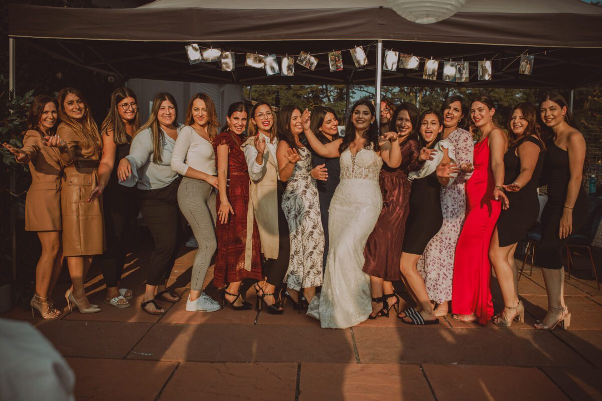 Eine Braut in einem weißen Brautkleid steht in der Mitte einer Gruppe lächelnder und untergehakter Frauen. Die Frauen, die in unterschiedlichen Outfits gekleidet sind, stehen unter einem Baldachin, der mit Lichterketten und hängenden Fotos geschmückt ist. Die Kulisse scheint eine Feier im Freien zu sein, die die Freude ihres Kennenlernens einfängt. - Elis Wedding - Hochzeitsfotograf Frankfurt