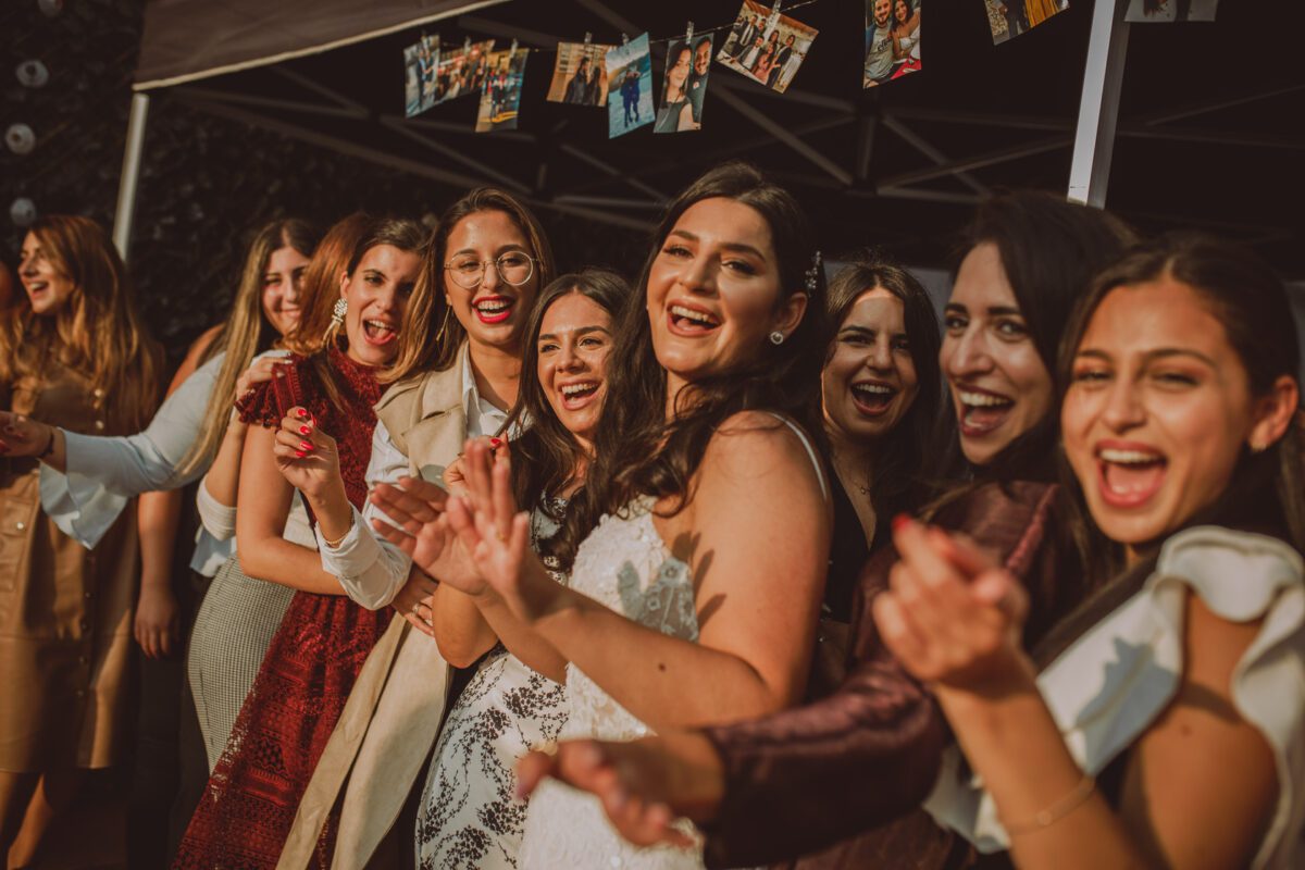 Eine Gruppe von Frauen steht eng beieinander unter einem Baldachin, lächelt und klatscht freudig während der standesamtlichen Trauung. Sie tragen eine Vielzahl von Outfits, von formellen Kleidern bis zu legeren Jacken. Über ihnen hängen Fotos ihres Hochzeitsfotografen auf einer Leine, was zur feierlichen Atmosphäre beiträgt. Das Licht ist warm und golden. - Elis Wedding - Hochzeitsfotograf Frankfurt