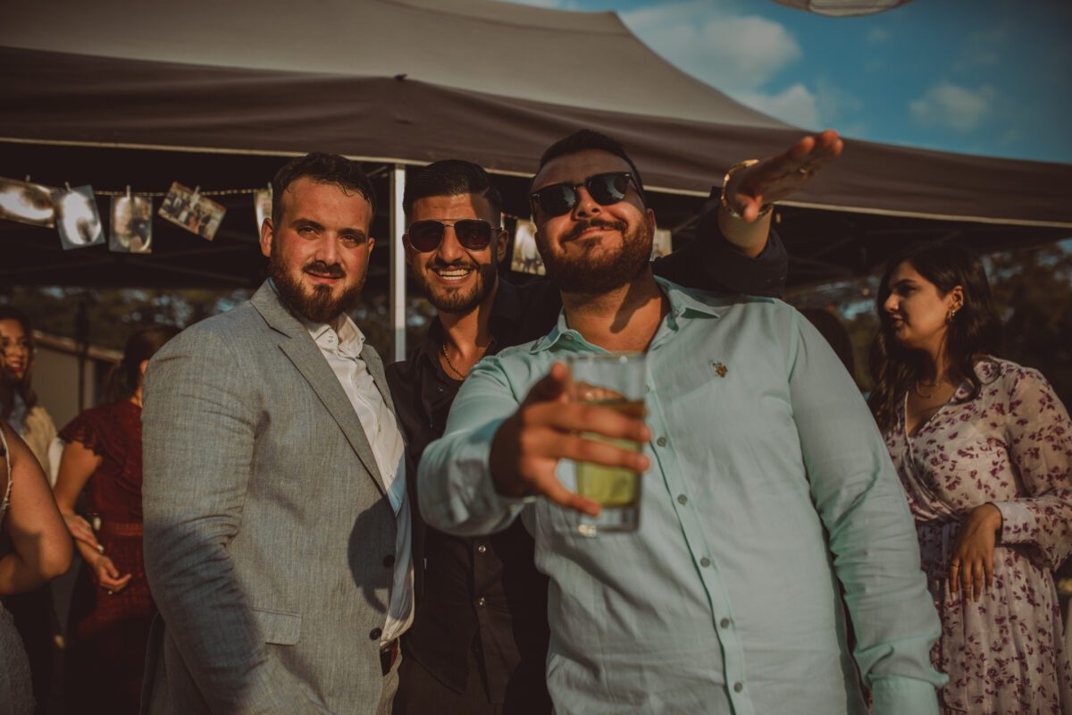 Drei Männer posieren für ein Foto während einer Outdoor-Veranstaltung unter einem Zelt. Der Mann in der Mitte trägt eine Sonnenbrille und ein schwarzes Outfit, während die Männer neben ihm helle Hemden und Blazer tragen. Der Mann rechts hält ein Getränk und streckt seinen anderen Arm nach vorne, um Erinnerungen an dieses Fotoshooting festzuhalten. Im Hintergrund sind Menschen und Fotos zu sehen. - Elis Wedding - Hochzeitsfotograf Frankfurt