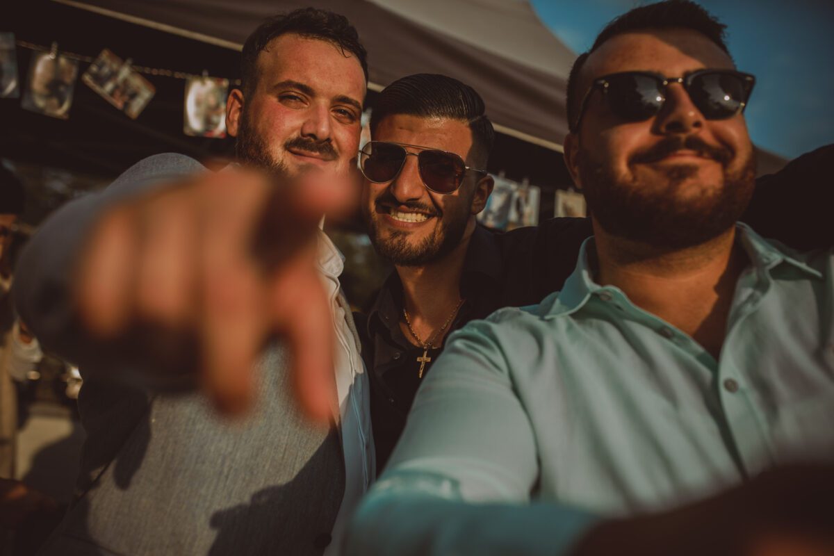 Drei Männer posieren gemeinsam bei einer Open-Air-Veranstaltung in Frankfurt. Der Mann links zeigt lächelnd in die Kamera. Der Mann in der Mitte steht selbstbewusst da und trägt ein dunkles Hemd, eine Sonnenbrille und eine goldene Kreuzkette. Der Mann rechts trägt ein hellblaues Hemd und eine Sonnenbrille. Ein Zelt mit aufgehängten Fotos dient als Kulisse für ihren spontanen Kennenlern-Moment. - Elis Wedding - Hochzeitsfotograf Frankfurt